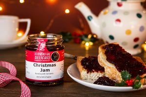 An open jar of Cumbrian Delights Christmas Jam sitting on a wooden table, next to a plate of toast spread with the rich purple jam